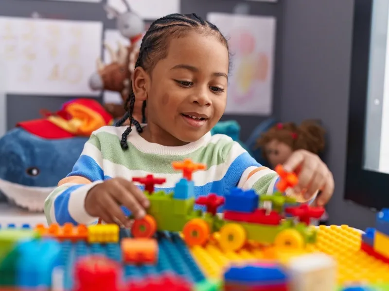 A young child with braided hair smiling while building with bright plastic blocks at a table in a sunny playroom