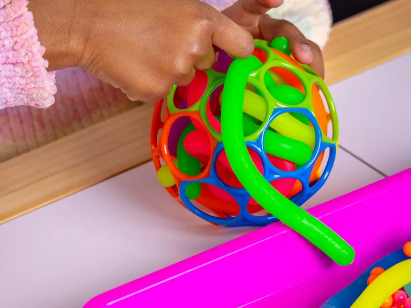 Close-up of a young child&rsquo;s hands gripping a brightly colored open-frame plastic ball and a bendy green tube on a table