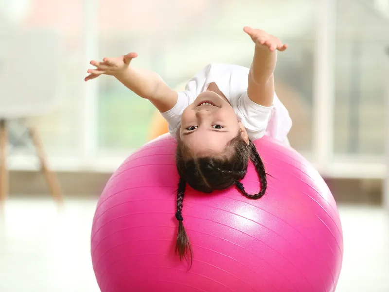 A smiling young girl lying backward over a large pink exercise ball with both arms stretched out overhead