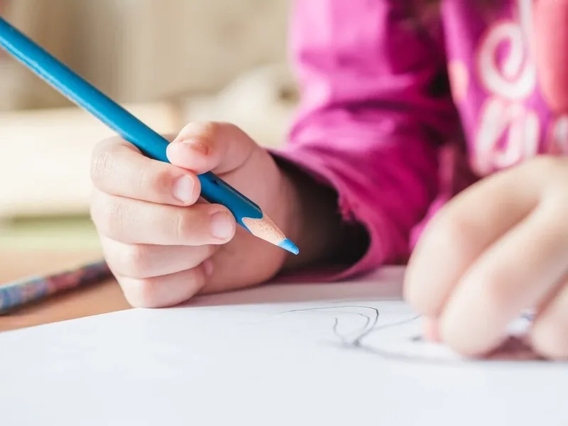 Close-up of a child&rsquo;s hand gripping a blue colored pencil and drawing a curved line on white paper