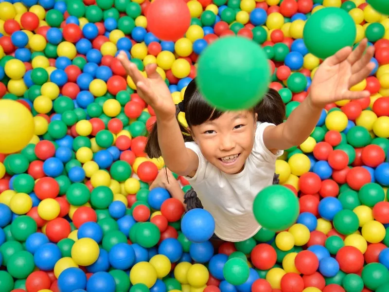 A smiling young girl lying in a ball pit full of red, yellow, blue and green plastic balls, reaching up with both hands