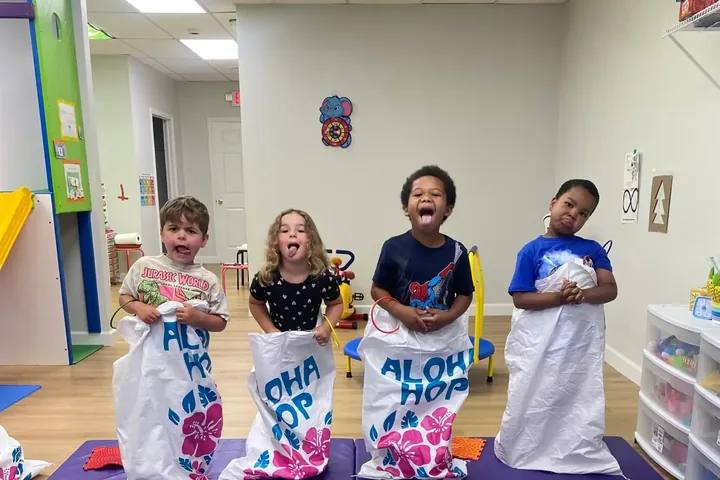 Four children standing in sack-race bags on a purple mat in the Planet Sensory gym in Baldwin, tongues out, with a yellow slide and storage bins behind them