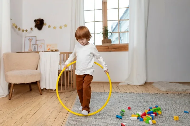 A young boy holding a yellow hula hoop around his waist and stepping through it, with building blocks scattered on the rug behind him