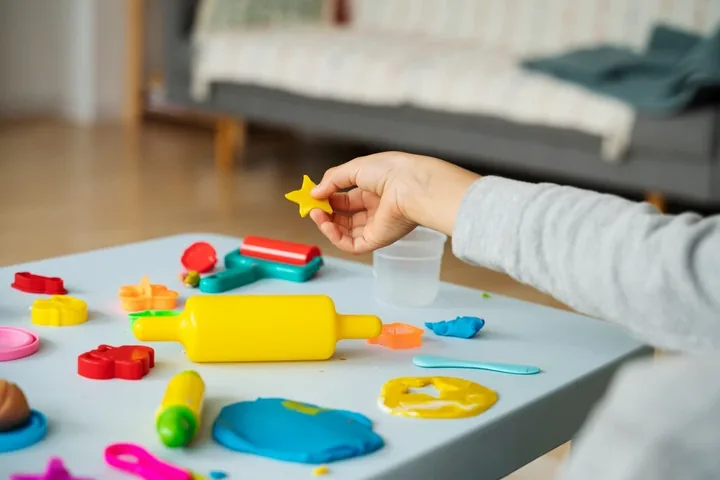 A child&rsquo;s hand lifting a small yellow play dough star off a table covered in cutters, a rolling pin and blue and yellow dough