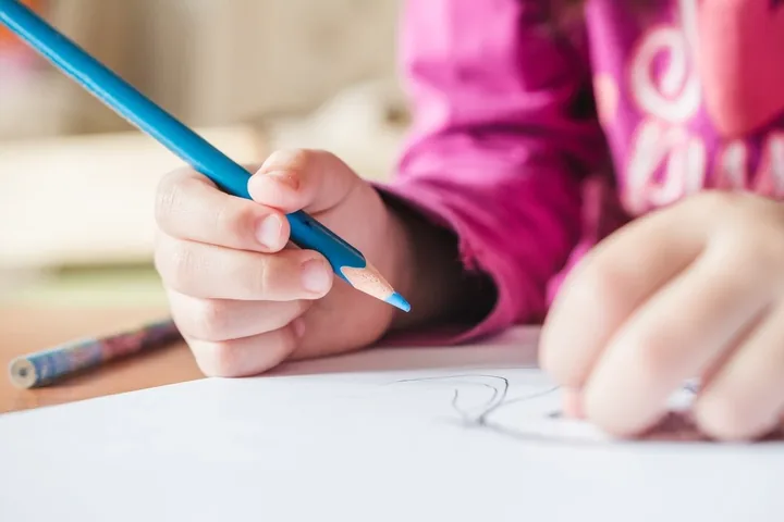 Close-up of a girl in a pink sweatshirt gripping a blue colored pencil and drawing on a sheet of white paper