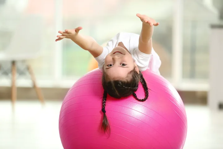 A girl lying back over a large pink therapy ball with both arms stretched overhead, grinning upside down