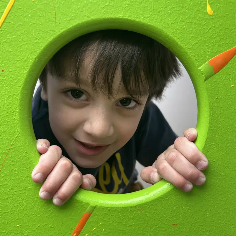 A smiling boy peeking through a round hole in a bright green play wall at the Planet Sensory gym in Baldwin