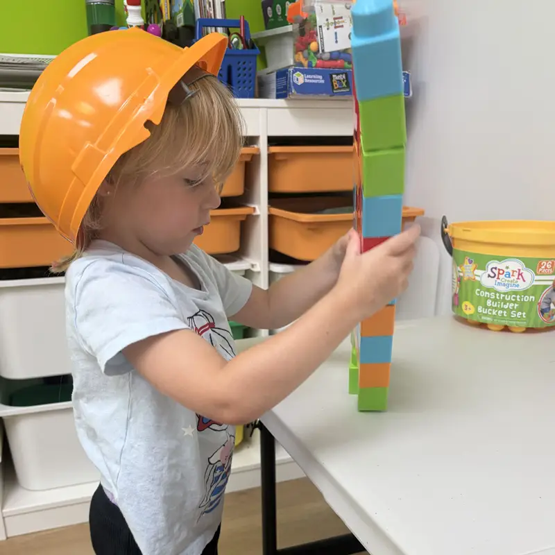 A young child in an orange toy hard hat carefully stacking a tall tower of colorful building blocks