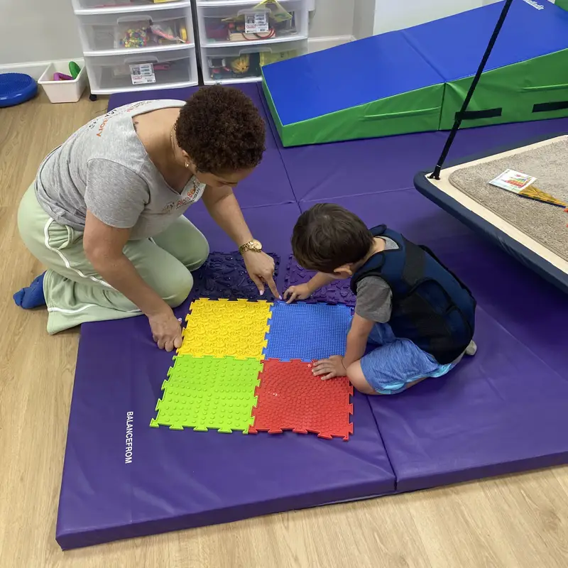 A therapist kneeling on a purple mat with a young boy, pointing to colorful textured sensory tiles