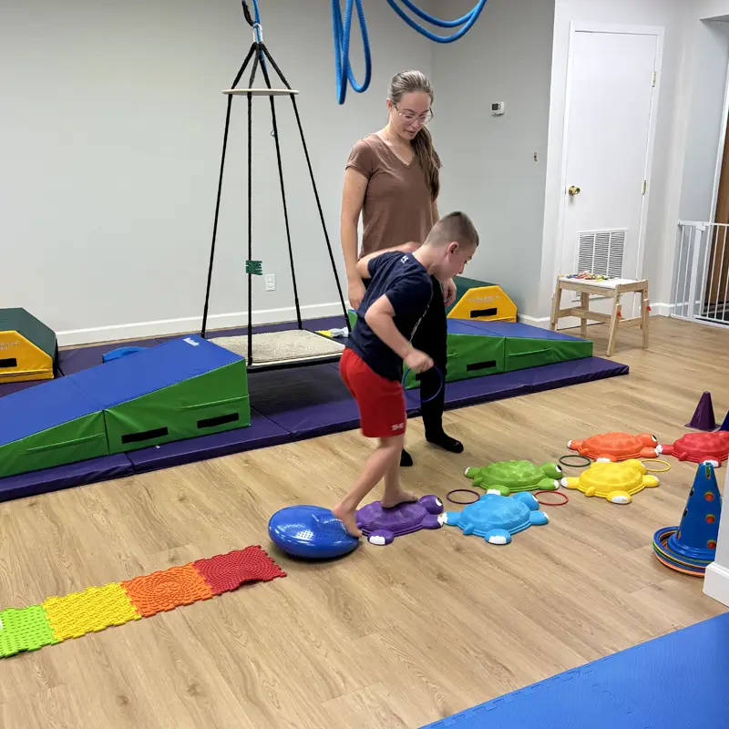 A boy stepping across balance stones in the sensory gym while a therapist stands beside him