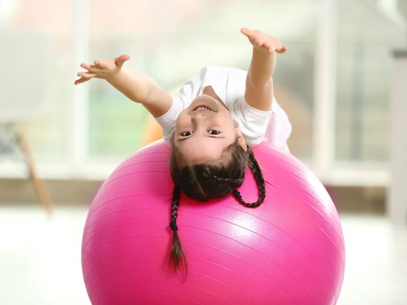 A young girl leaning back over a pink exercise ball with both arms raised