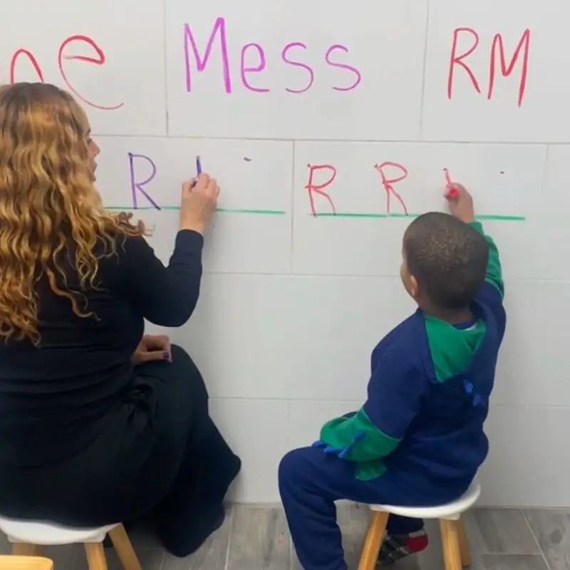 An occupational therapist and a young boy sitting on stools, writing letters side by side on a whiteboard wall