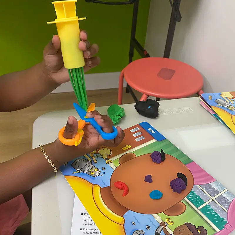 A child using a play-dough extruder and bright plastic scissors over a feelings mat at a therapy table