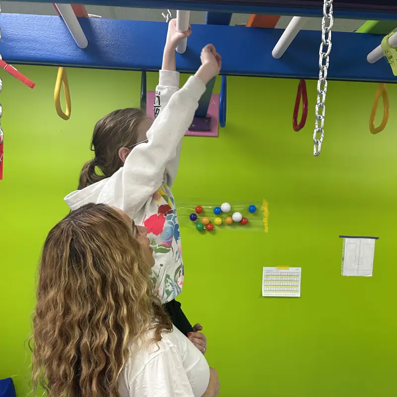 A therapist helping a girl reach up to grab the monkey bars in the sensory gym