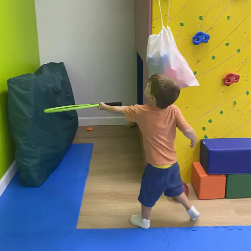 A boy reaching a green hoop toward a crash pad beside the yellow climbing wall