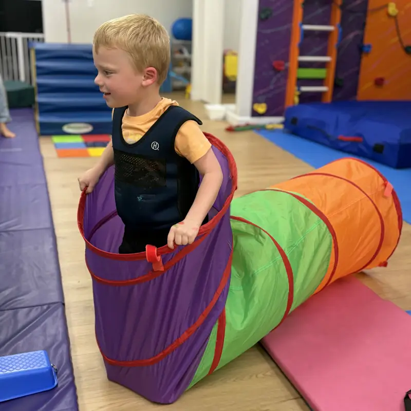 A smiling boy in a weighted vest climbing out of the end of a colorful play tunnel
